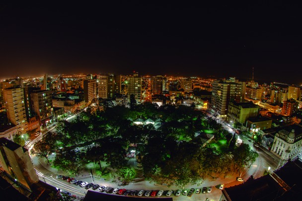 Fotografia da cidade de Pelotas de noite. No meio da foto, está uma praça iluminada nas extremidades e em sua volta, muitos prédios altos iluminados pelas luzes noturnas. 