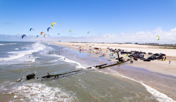 Parte da praia do Cassino em que aparece restos de uma embarcação encalhada. O céu está azul, com poucas nuvens e há pessoas se banhando na água, além de carros na areia da praia. 