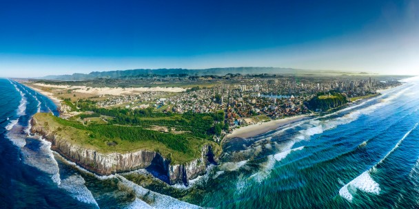 Foto do alto da cidade de torres, com céu azul, água cristalina. No meio da foto, está parte do morro da cidade, ao lado, a praia. Prédios estampam a parte urbana da cidade acima da praia. 