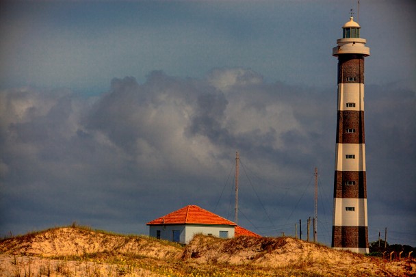 Farol de Mostardas, ao lado de uma pequena casa com telhado laranja. Céu azul escuro. 