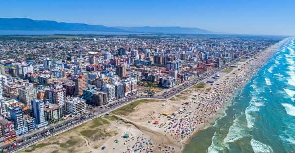 Praia de Capão da Canos vista de cima. À esquerda, prédios que compõem a cidade e a direita, a praia e o mar. 