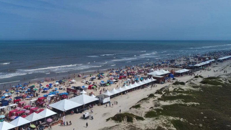 Praia de Xangri-Lá vista de cima. Na areia, tem toldos e guarda-sóis de diversas cores por toda a extensão da praia.