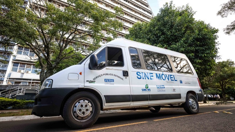 Mini van branca do Sine Móvel estacionada em frente ao prédio do Centro Administrativo Fernando Ferrari. Atrás do veículo, existem árvores com folhas verdes.