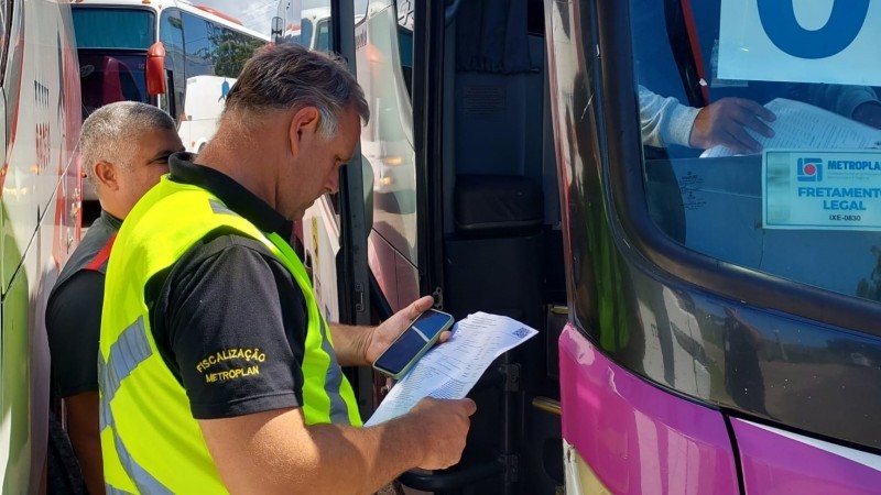 Dois homens em frente a um ônibus. Um deles está lendo um papel e o outro está olhando para frente. 