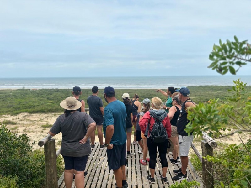Um pequeno grupo de pessoas em cima de uma plataforma de madeira, em frente ao mar. O grupo tem adultos, homens e mulheres, que estão usando roupas esportivas e chapéus e bonés.