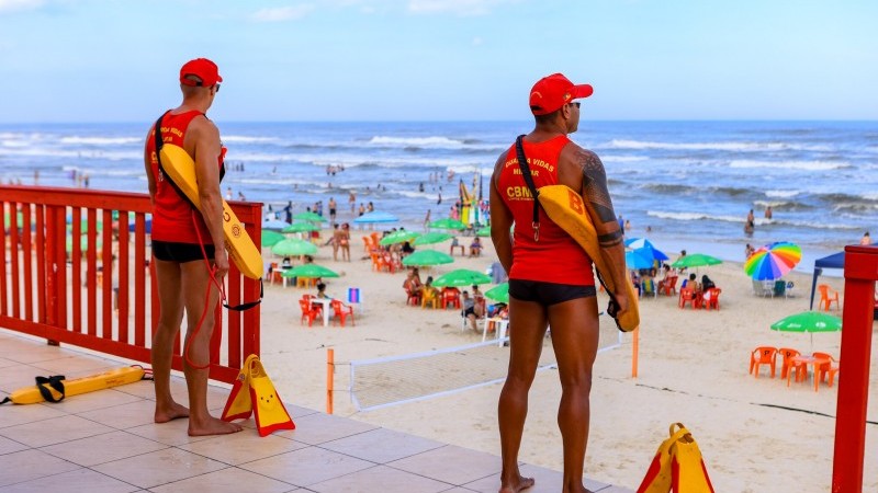 Dois guarda-vidas posicionados de costas observam o movimento de banhistas em praia