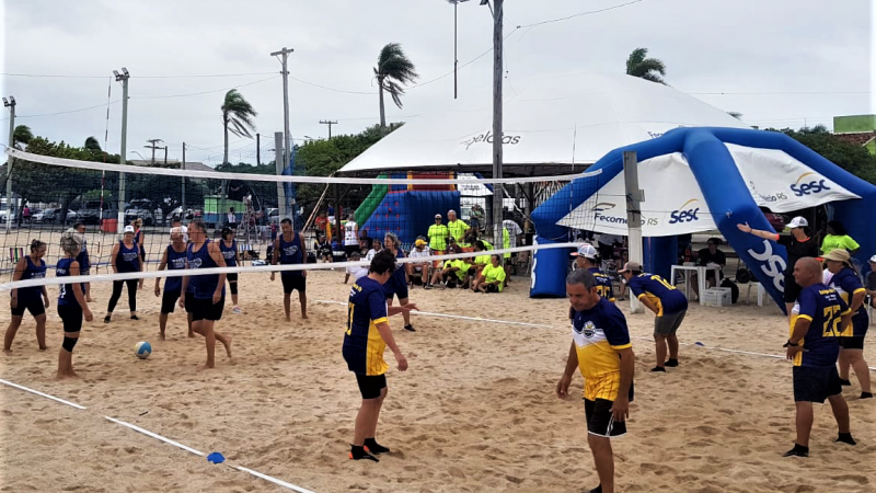 Grupo de pessoas, com homens e mulheres jogando vôlei em uma praia. Eles usam roupas amarelas e azul marinho, ao lado de um tenda do Sesc. O céu está um pouco nublado, com algumas nuvens e sem sol. 