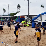 Grupo de pessoas, com homens e mulheres jogando vôlei em uma praia. Eles usam roupas amarelas e azul marinho, ao lado de um tenda do Sesc. O céu está um pouco nublado, com algumas nuvens e sem sol. 