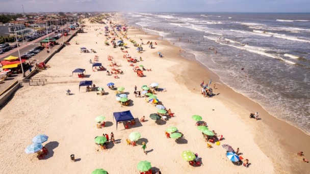 Vista de cima de uma praia. Na areia, há guarda-sóis verdes e coloridos. Algumas pessoas estão no mar. Alguns carros estão estacionados em frente ao calçadão da praia. 