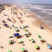 Vista de cima de uma praia. Na areia, há guarda-sóis verdes e coloridos. Algumas pessoas estão no mar. Alguns carros estão estacionados em frente ao calçadão da praia. 