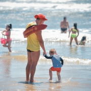 A imagem mostra um guarda-vida de costas, segurando a mão de uma criança. Ele está de frente para o mar, onde três crianças e um adulto se banham.