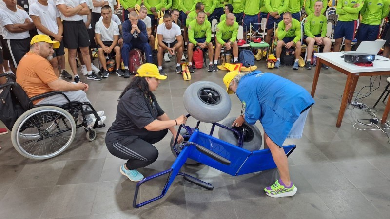 A fotografia mostra as coordendoras da Faders, do Rede praia acessível e de Acessibilidade, usando bonés amarelos do Verão Total enquanto demonstram, no chão, o funcionamento de uma cadeira anfíbia azul, utilizada no Programa Praia Acessível. Uma delas está agachada e a outra está inclinada para frente, ajustando uma das rodas grandes do equipamento. Ao lado esquerdo da imagem, o presdiente da Faders sentado em cadeira de rodas observa atentamente a demonstração. Ao fundo, diversos guardas-vidas civis e militares, alguns com camiseta branca e outros com camiseta verde, acompanham o treinamento sentados e em pé. À direita, há uma mesa com projetor ligado e cabos no chão, reforçando o ambiente de capacitação.