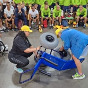 A fotografia mostra as coordendoras da Faders, do Rede praia acessível e de Acessibilidade, usando bonés amarelos do Verão Total enquanto demonstram, no chão, o funcionamento de uma cadeira anfíbia azul, utilizada no Programa Praia Acessível. Uma delas está agachada e a outra está inclinada para frente, ajustando uma das rodas grandes do equipamento. Ao lado esquerdo da imagem, o presdiente da Faders sentado em cadeira de rodas observa atentamente a demonstração. Ao fundo, diversos guardas-vidas civis e militares, alguns com camiseta branca e outros com camiseta verde, acompanham o treinamento sentados e em pé. À direita, há uma mesa com projetor ligado e cabos no chão, reforçando o ambiente de capacitação.