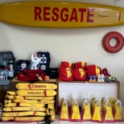A imagem mostra uma sala com diversos equipamentos de salvamento utilizados por guarda-vidas. Na parede, h&aacute; uma prancha de resgate amarela com a palavra &ldquo;RESGATE&rdquo; em vermelho. Ao lado, est&aacute; pendido um flutuador circular vermelho. Sobre uma mesa, encontram-se mochilas vermelhas, bolsas de primeiros socorros, cilindros de oxig&ecirc;nio, colar cervical, protetor solar e outros itens m&eacute;dicos. No ch&atilde;o, empilhados, h&aacute; v&aacute;rios flutuadores amarelos com a inscri&ccedil;&atilde;o &ldquo;CBMRS &ndash; GUARDA-VIDAS&rdquo;, al&eacute;m de pares de nadadeiras tamb&eacute;m nas cores amarela e vermelha embaladas em pl&aacute;stico. &Agrave; esquerda, aparecem p&aacute;s e outros materiais de apoio.