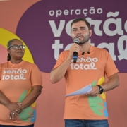 Uma foto em close-up, capturada em um evento interno, mostra um grupo de quatro pessoas paradas em frente a uma parede de fundo colorida que promove a "opera&ccedil;&atilde;o ver&atilde;o total".O foco principal est&aacute; em um homem de pele clara no centro-direita, que est&aacute; falando em um microfone que segura com a m&atilde;o direita. Ele veste uma camiseta laranja promocional com o logotipo "opera&ccedil;&atilde;o ver&atilde;o total" em letras brancas. Ele segura um peda&ccedil;o de papel dobrado na m&atilde;o esquerda e usa um rel&oacute;gio de pulso preto.Ao lado dele, no centro-esquerda, est&aacute; uma mulher de pele escura com cabelo curto e &oacute;culos. Ela tamb&eacute;m veste a mesma camiseta laranja promocional e tem os bra&ccedil;os cruzados na frente.Outros dois homens aparecem parcialmente:&Agrave; esquerda, um homem de pele clara com uma camiseta escura.&Agrave; extrema direita, um homem de pele clara em uma camisa cinza-escura.O fundo &eacute; um painel de parede com as palavras "opera&ccedil;&atilde;o ver&atilde;o total" escritas em branco em grandes faixas horizontais de cores laranja, roxo e verde-&aacute;gua. A faixa roxa, mais proeminente, cont&eacute;m a palavra "ver&atilde;o" e o til ($\tilde{}$) alongado.