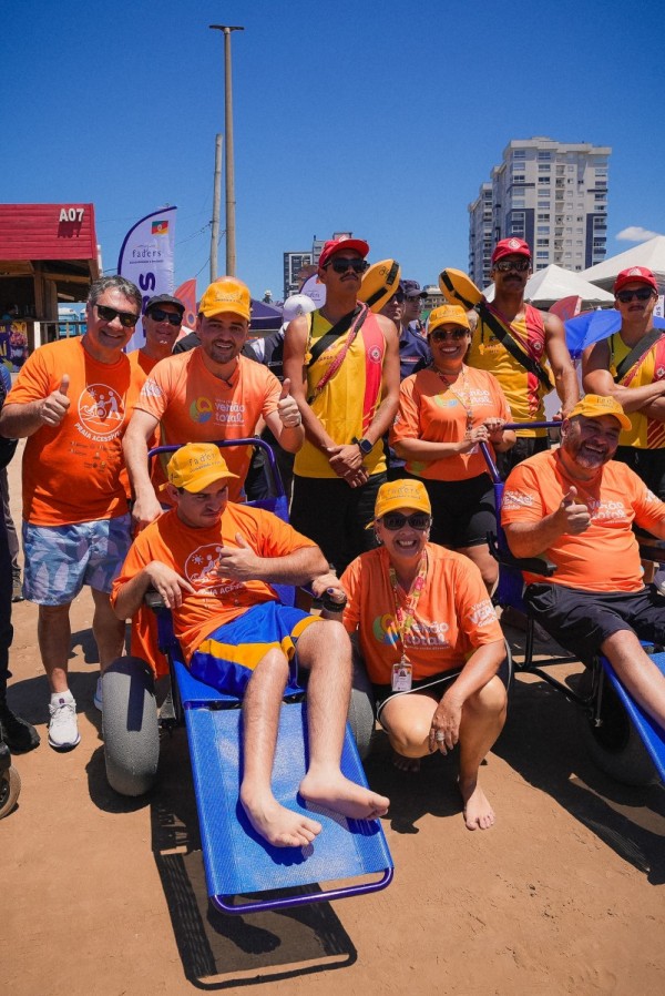 Foto mostra o vice-governador Gabriel Souza na Operação Verão Total com um cadeirante e outros servidores. Todos vestem camisetas laranjas.